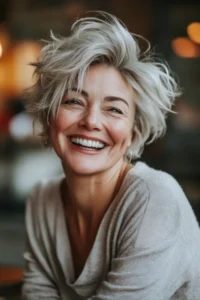 Effortless Shaggy Pixie with Movement. A woman over 50 with short, layered gray hair is smiling widely, wearing a light-colored sweater. The background is softly blurred.