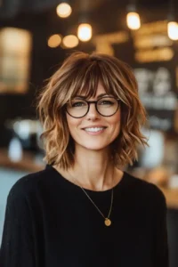 Modern Shaggy Bob with Side-Swept Fringe. A stylish woman over 40 with shoulder-length wavy hair and glasses smiles in a warmly lit, modern interior.